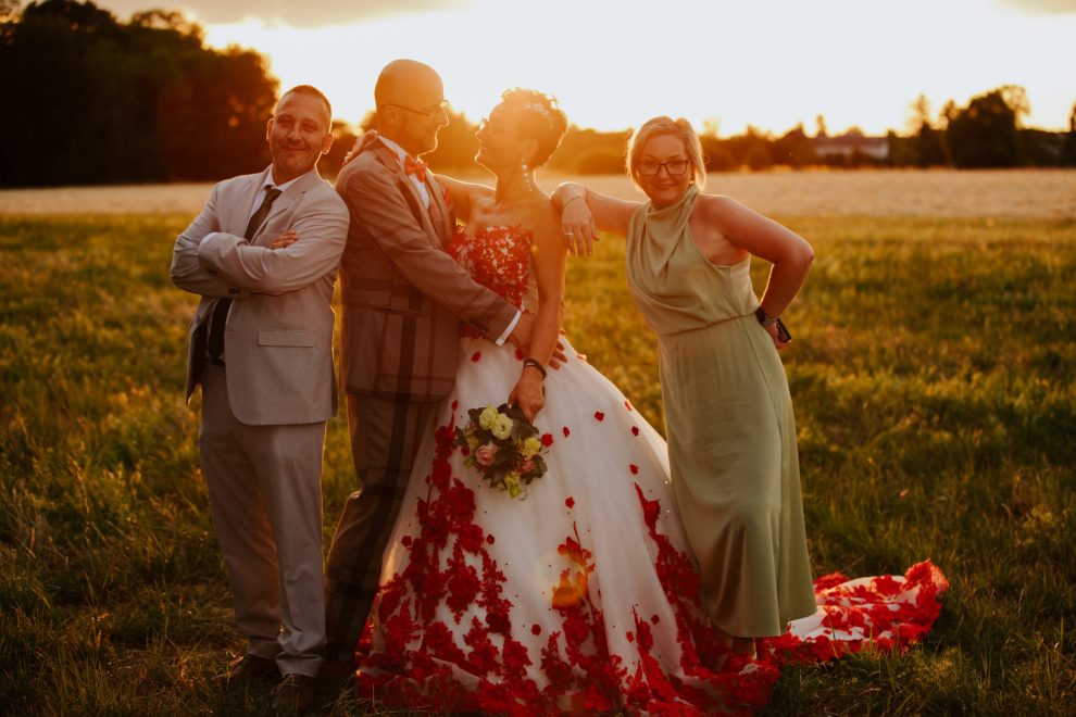 Romantischer Sonnenuntergang in der goldenen Stunde. Sonnenstrahlen scheinen in die Kamera. Am Waldrand und Feld stehen Braut und Bräutigam einander zugewandt. Der Bräutigam trägt einen grauen Old-School-Anzug mit Karomuster und hält seine Hände an der Taille der Braut. Die Braut trägt ein bodenlanges weißes Kleid mit roten Spitzenblumen. Ihre rechte Hand umfasst den Hals des Bräutigams, während sie in ihrer linken Hand einen Blumenstrauß hält. Der Trauzeuge steht lächelnd mit verschränkten Armen neben dem Bräutigam. Die Trauzeugin steht in entspannter Haltung neben der Braut und legt eine Hand auf deren Schulter. Beide Frauen lächeln.